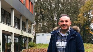 Davide Servello steht mit seiner dunjelblauen jacke draußen vor einem Gebäude. (Foto: UNSERDING)