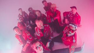 Alle elf Bandmitglieder sitzen in roter Jacke im klassischen Marching Band-Look auf dem Boden unn gucken in die Kamera, die Kamera blickt von oben auf die Band runter. (Foto: Atelier (Presse))